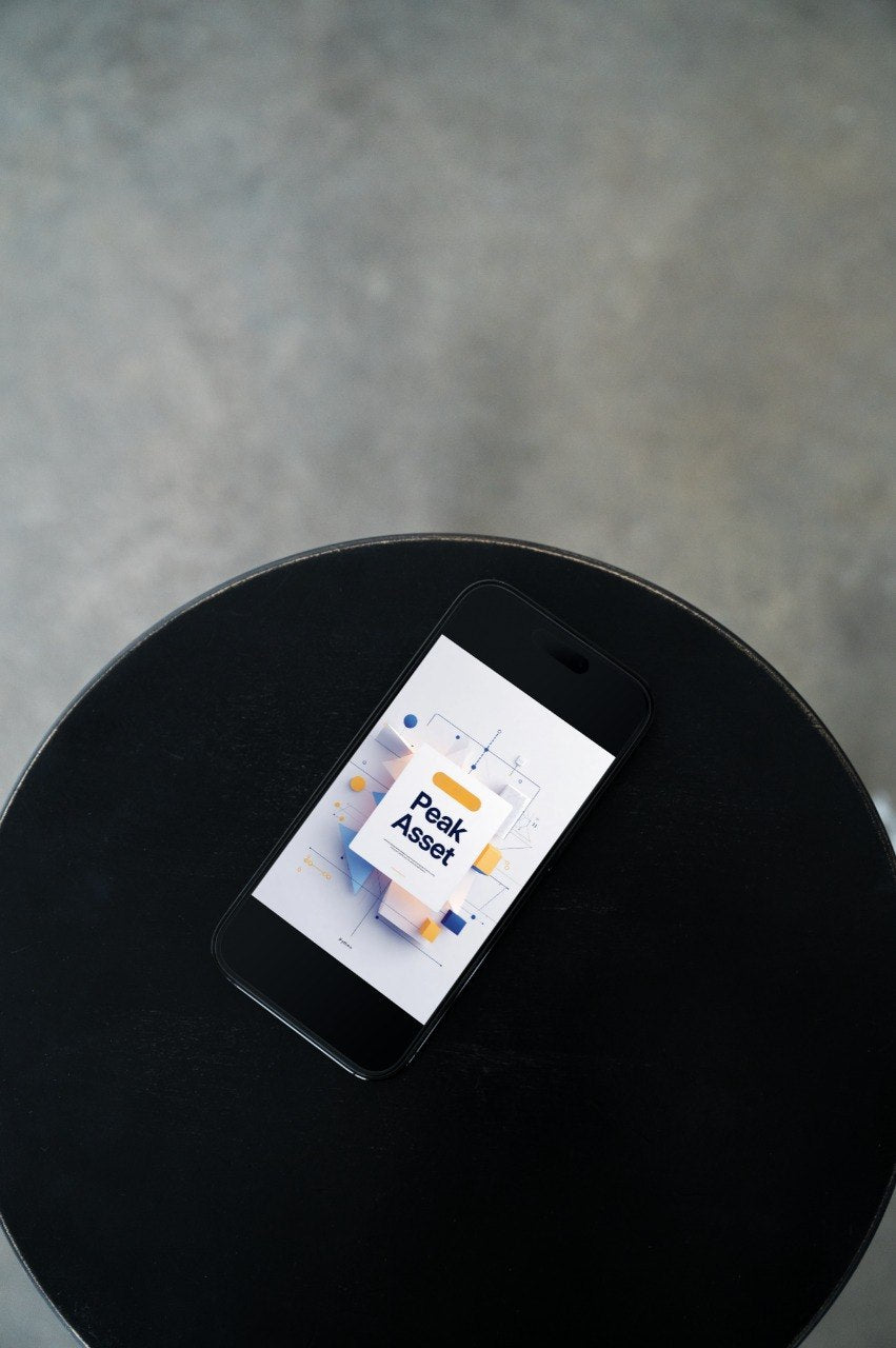 A black smartphone lying on a round black table, displaying a digital interface with the text 'Peak Asset'. The background is a textured gray concrete floor.